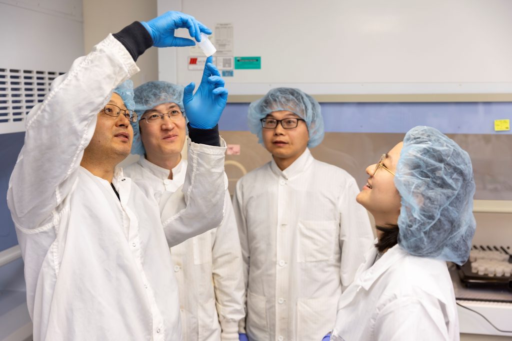 Four scientists in white lab suits looking at a glass vial. In a white (clean) lab.