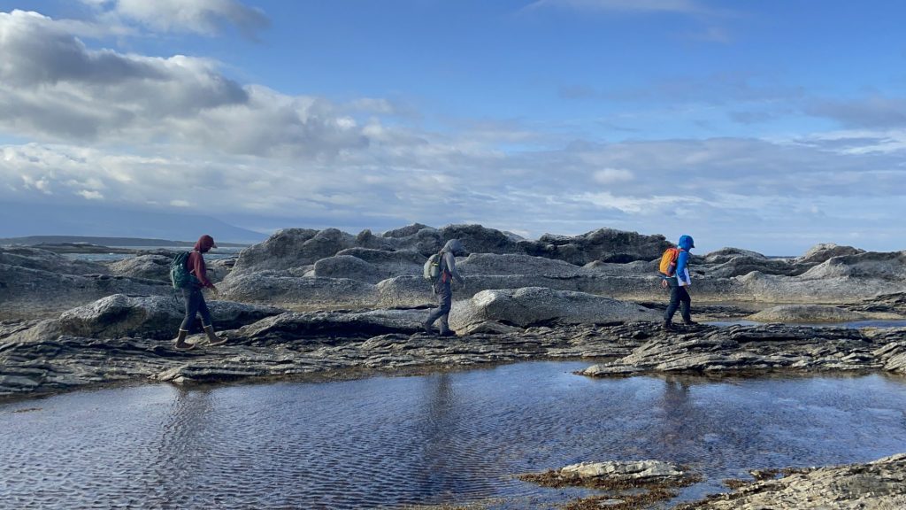 Three people hiking in front of a stream, with rocks and blue sky behind them, doing field work.
