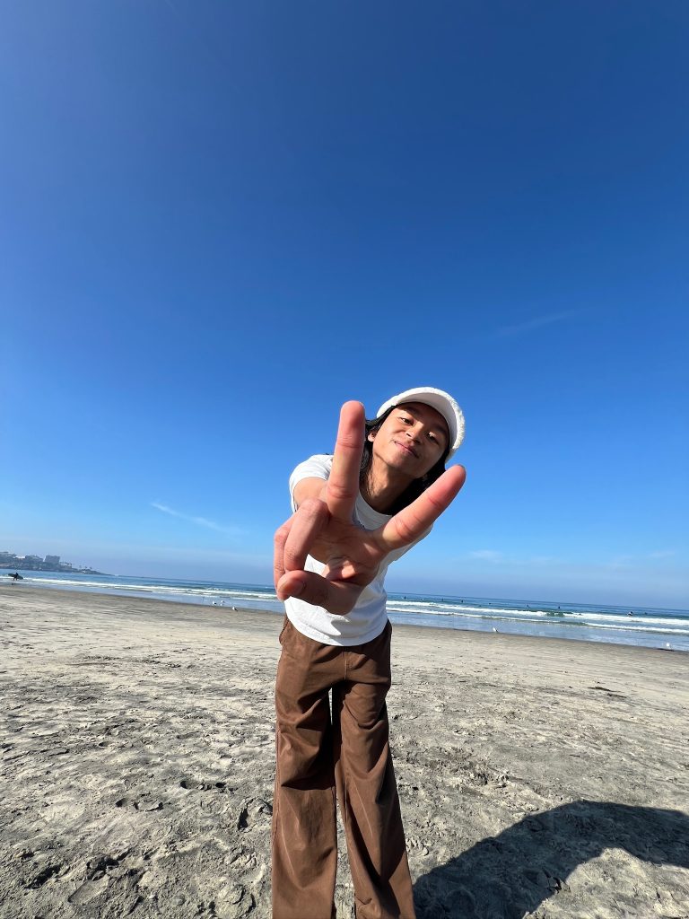 Close up of Fikri Roslan holding two fingers up to the camera, standing on the beach with the ocean in the background.