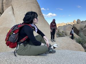 Student with medium dark hair, black longsleeve shirt, and green hiking pants sitting on light color rock with large rocks and other students in the background.