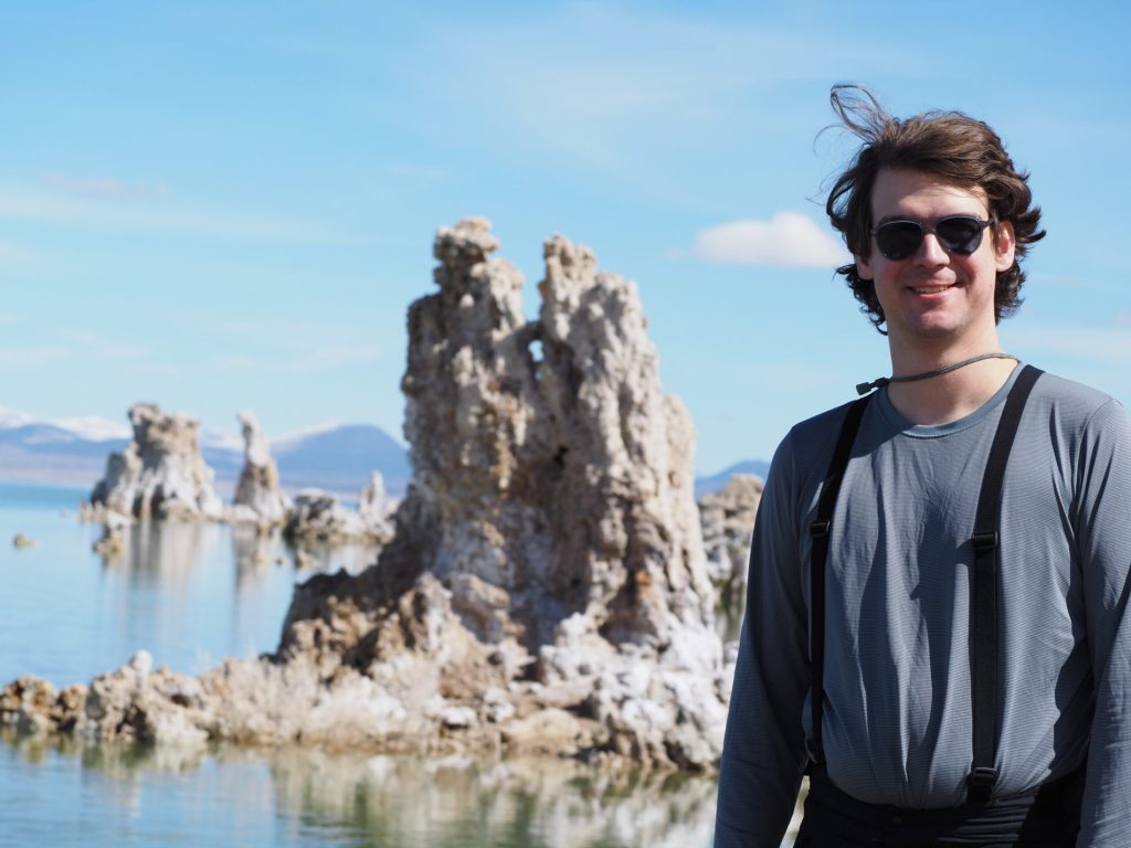 Student with short brown hair and grey long sleeve shirt with blurry background tall rocks sticking out of water, blue skies, and mountains in the far distance.