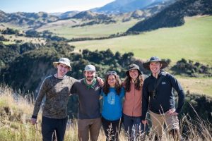 Five people smiling and posing for the camera, standing in a field with mountainous landscape in the background.