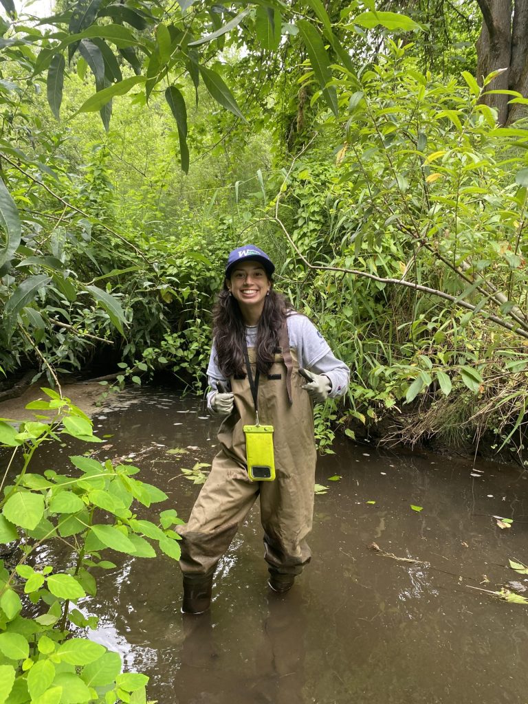 Grace Nice standing in a muddy stream with green foliage in the back ground.