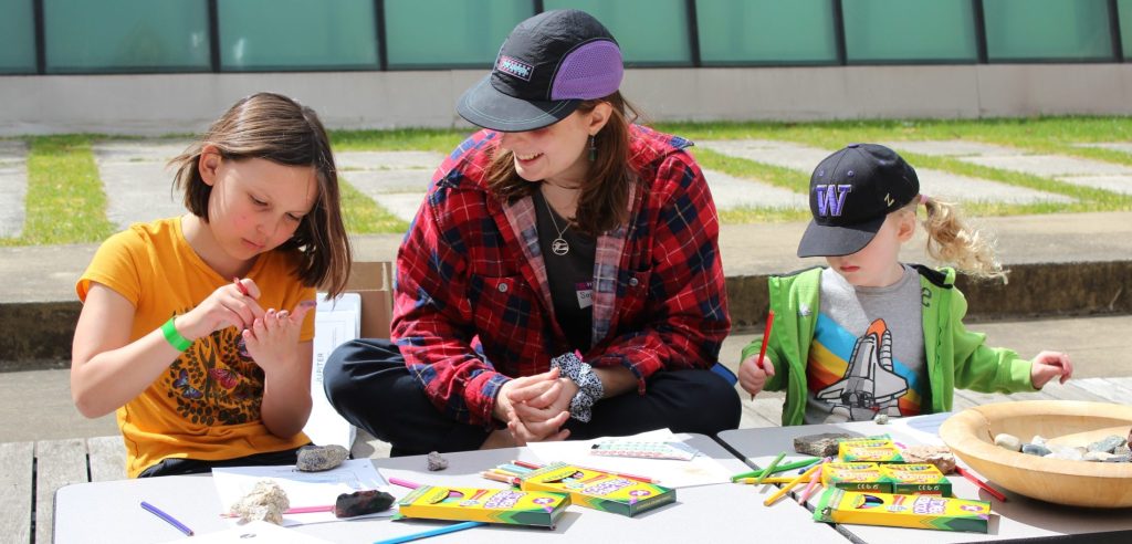 Two kids with a graduate student at the coloring table, working on coloring in geoscience-themed coloring pages.