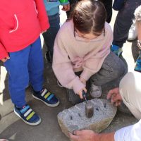 A young kid wearing safety glasses holds a small sledgehammer to hammer a row of pegs into a large rock to split it down the middle.