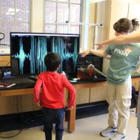 A young person jumps up and down, with the jumps recorded by a seismometer hooked up to a screen. The jumps are displayed on the screen as spikes.