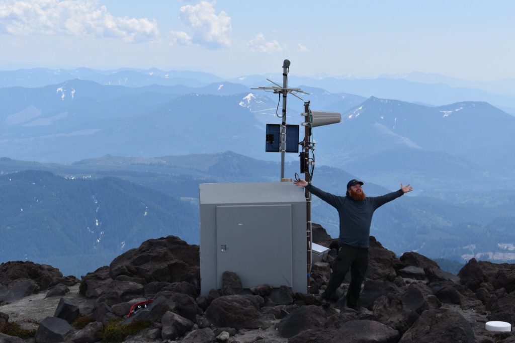 PNSN Field Engineer Doug Gibbons standing next to a seismology installation at Mount St. Helens with blue mountain ridges behind him.