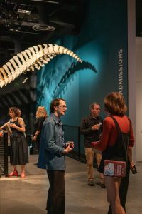 Department community members socializing at the ESS 130th Anniversary Celebration in the Burke Museum in front of a dinosaur fossil.