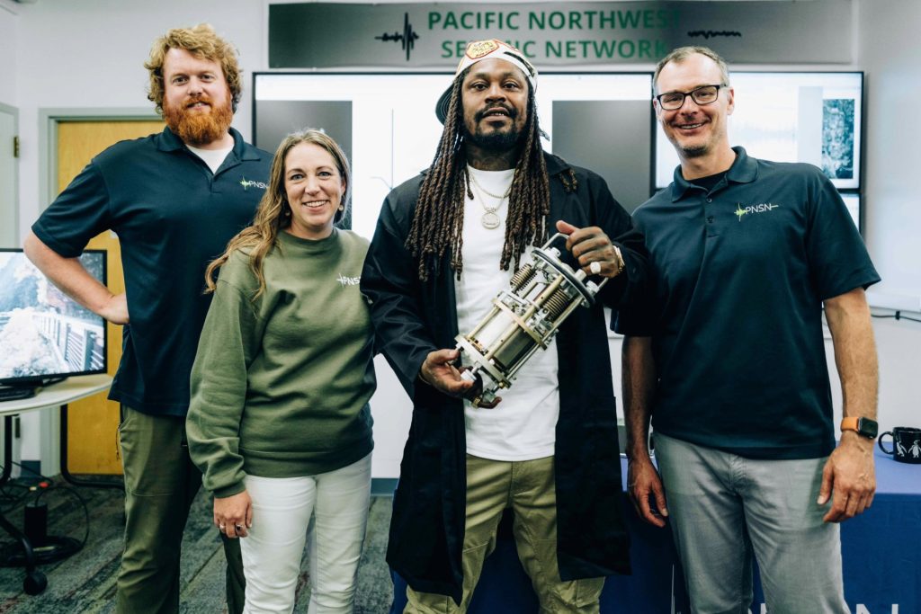 Group photo of PNSN staff Doug Gibbons, Amy Wright, and Alex Hutko standing around former Seahawk player Marshawn Lynch in front of a Pacific Northwest Seismic Network sign.