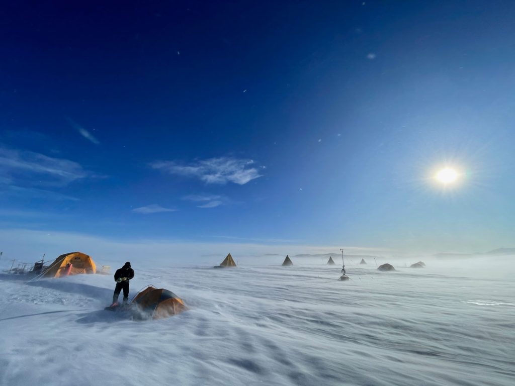 A windswept snowfield with a few people near small tents against a bright blue sky in Antarctica.