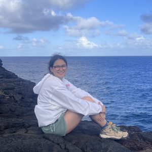 Close up of Lauren Kafadarian sitting on a black rock in front of the ocean.