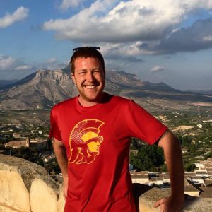Close up of Pete Wynn leaning against a wall with a mountain and town in the background.