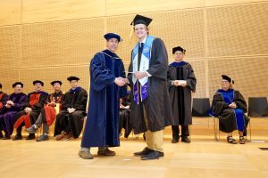 Professor and Chair David Schmidt and undergraduate in cap and gown shaking hands on stage.