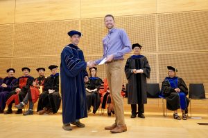 Professor and Chair David Schmidt and undergraduate in cap and gown shaking hands on stage.