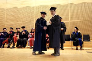 Professor and Chair David Schmidt and undergraduate in cap and gown shaking hands on stage.