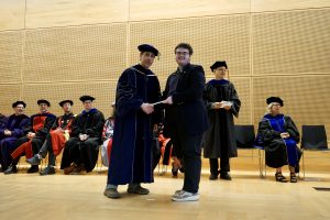 Professor and Chair David Schmidt and undergraduate in cap and gown shaking hands on stage.