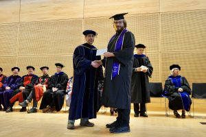 Professor and Chair David Schmidt and undergraduate in cap and gown shaking hands on stage.
