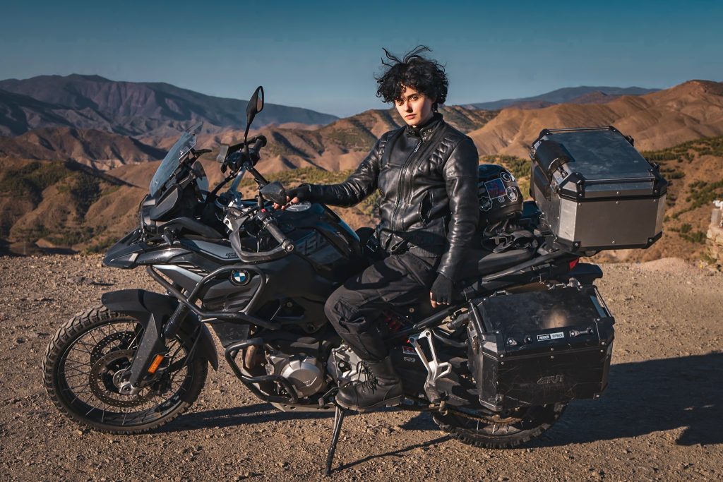 Pine Dysart-Bricken sitting on a black motorcycle in all black clothing in Morocco with mountains in the background.