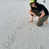 Autum Downey sitting on a beach pointing to an equation drawn in the sand.